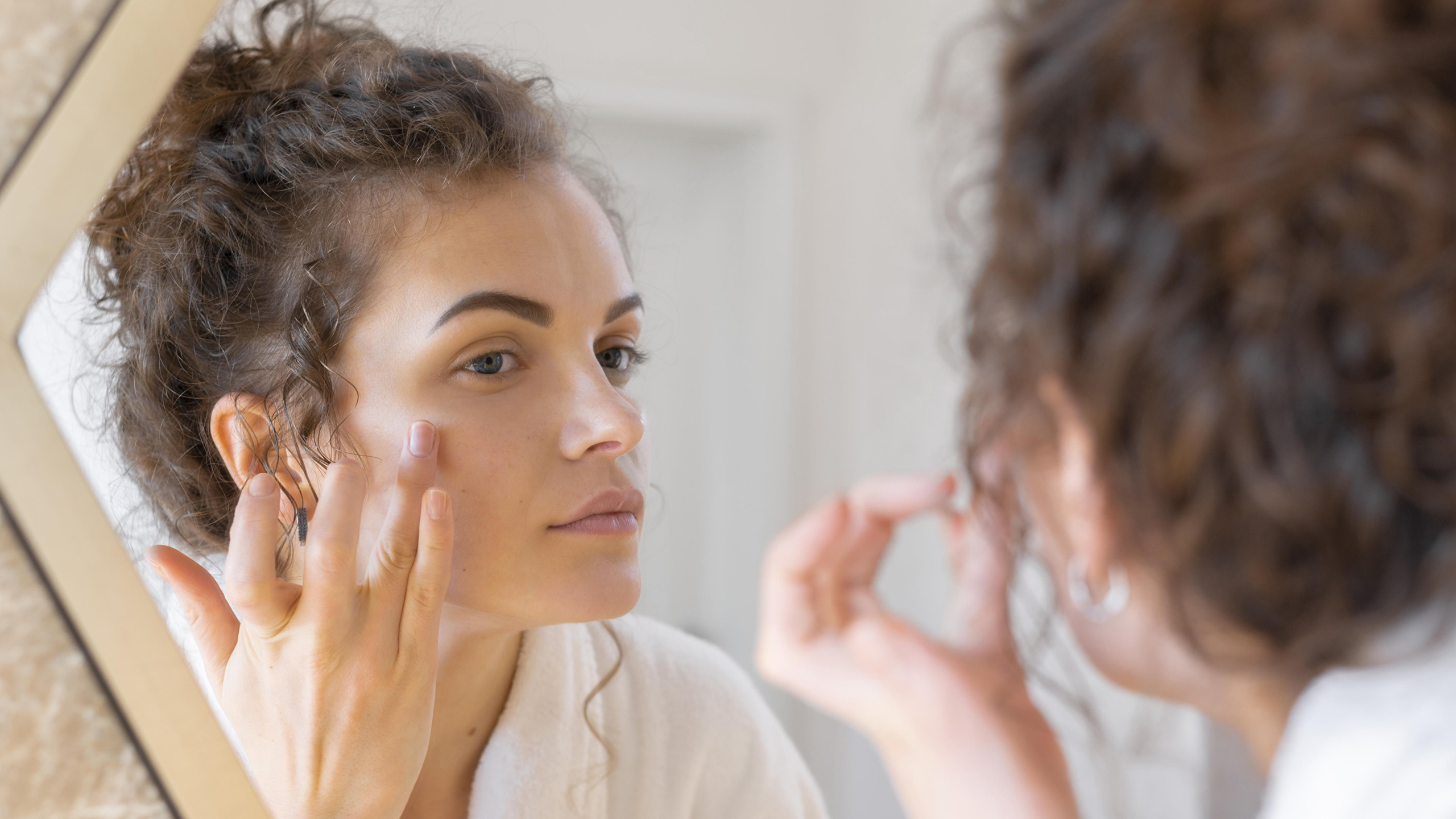 Woman looking in the mirror while gently touching her face during a skincare routine, representing common skin concerns like redness or irritation.