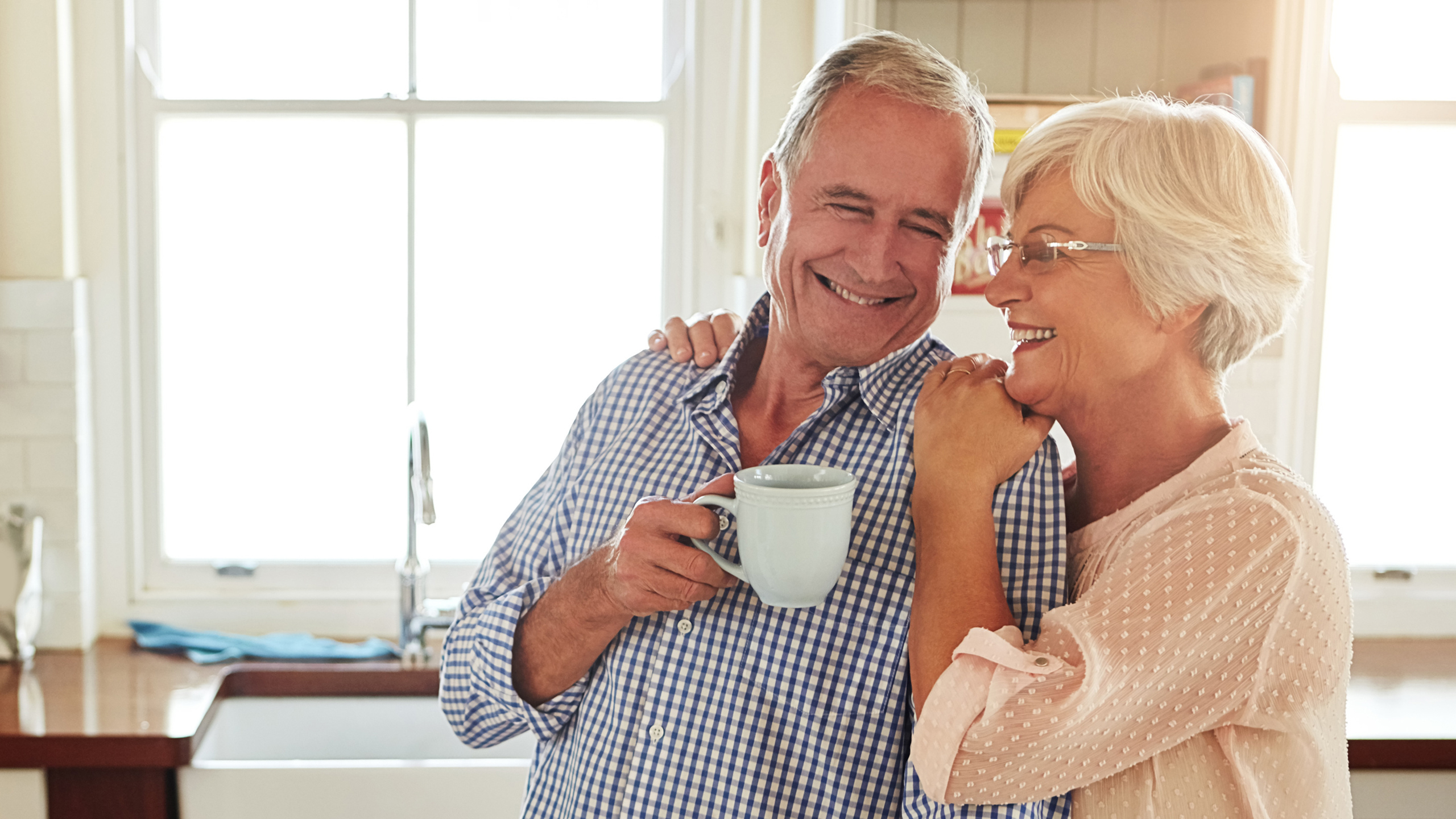 A mature couple sharing a warm morning hug in their kitchen while holding coffee cups.