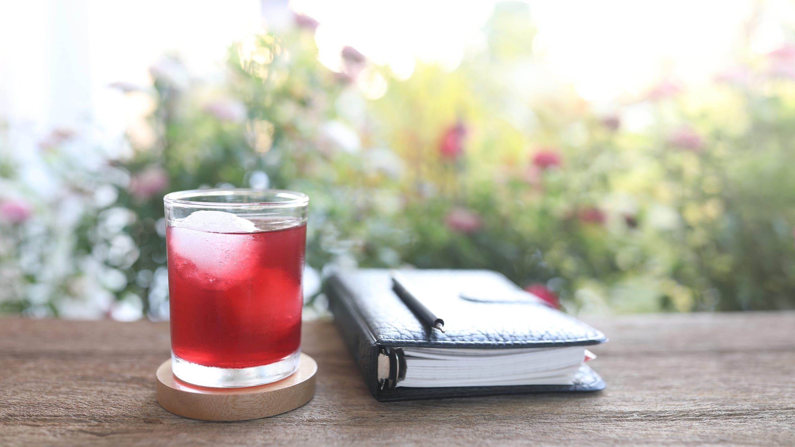 A pomegranate Unimate drink beside an open notebook and a daisy flower on a wooden table in natural outdoor light.