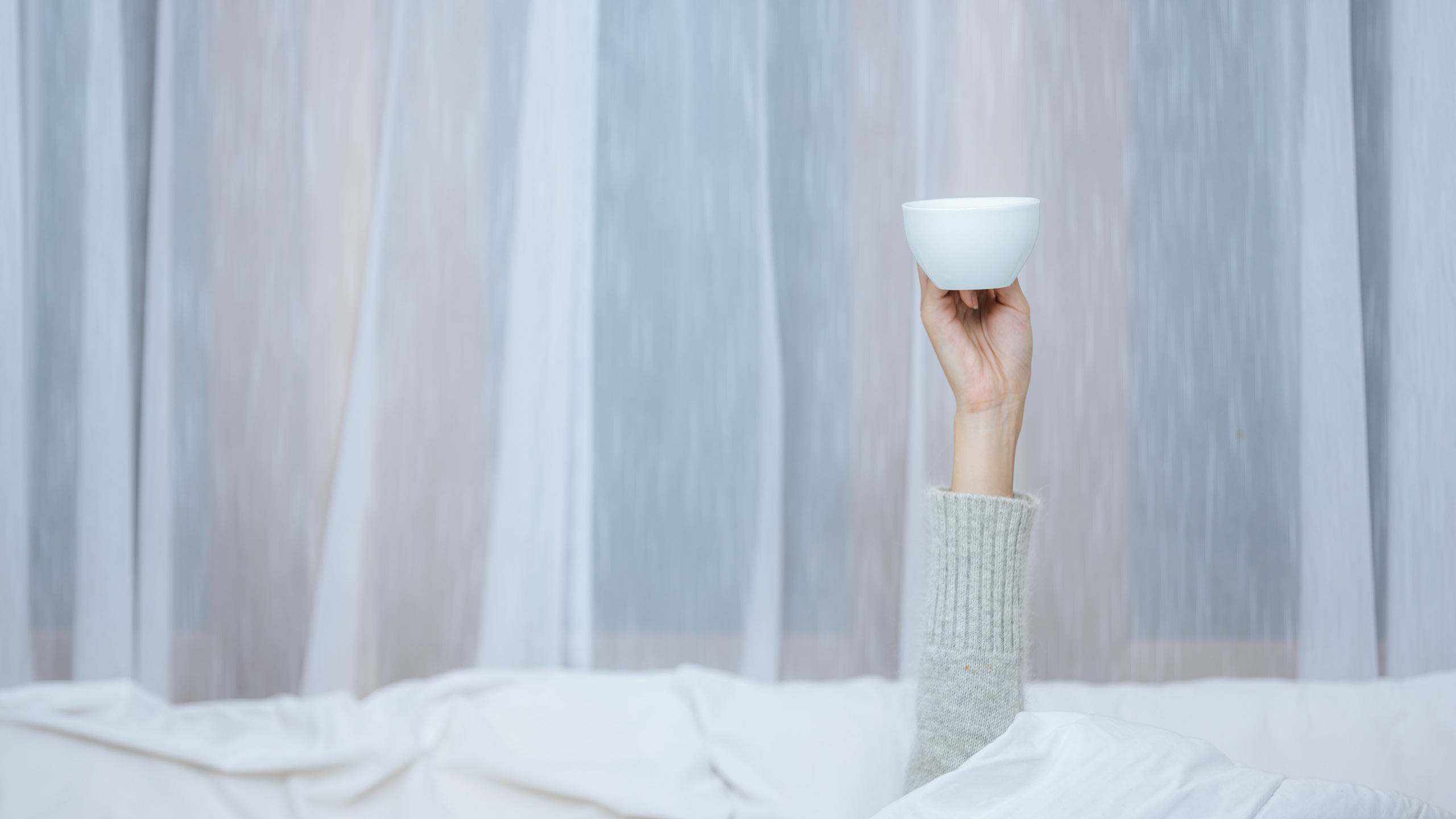 Woman lying back in bed holding a coffee mug, wrapped in a duvet in soft morning light